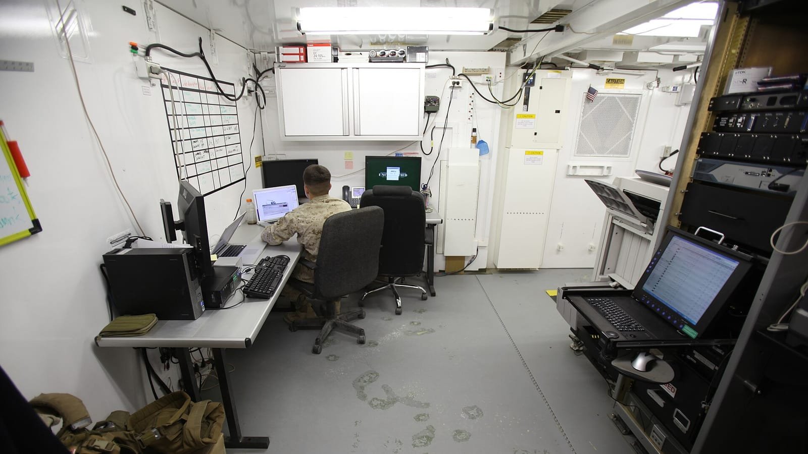 Un videógrafo del Cuerpo de Marines de EE. UU., visto de espaldas, edita vídeo en un portátil dentro de una cabina de producción con monitores y un rack de equipos