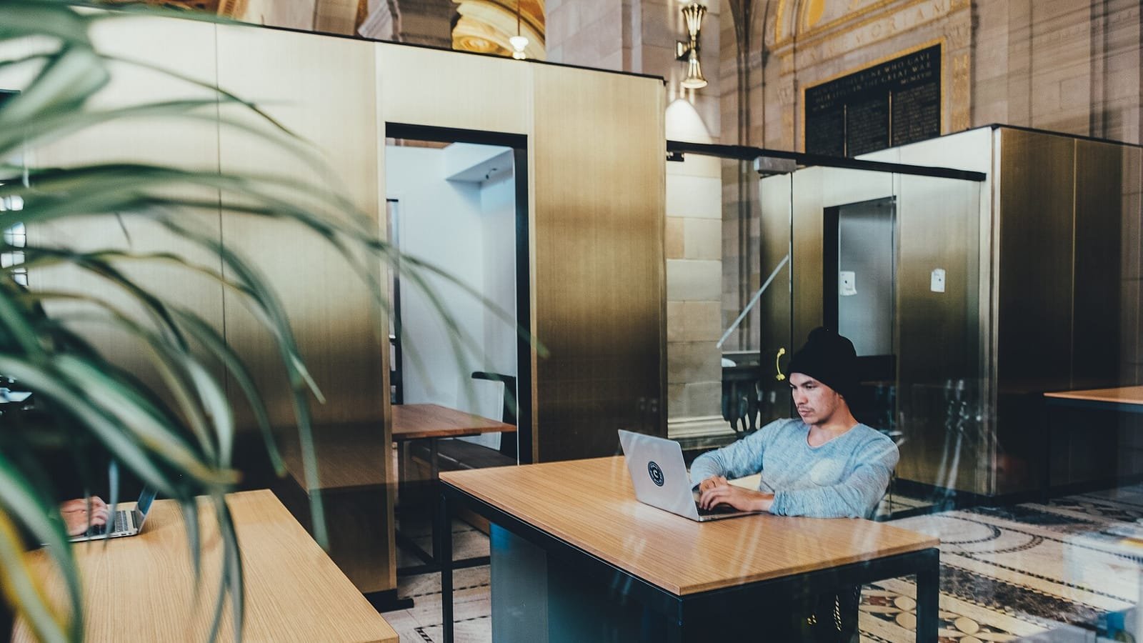 Un hombre con gorro trabaja con un portátil en una mesa de madera de un espacio de coworking