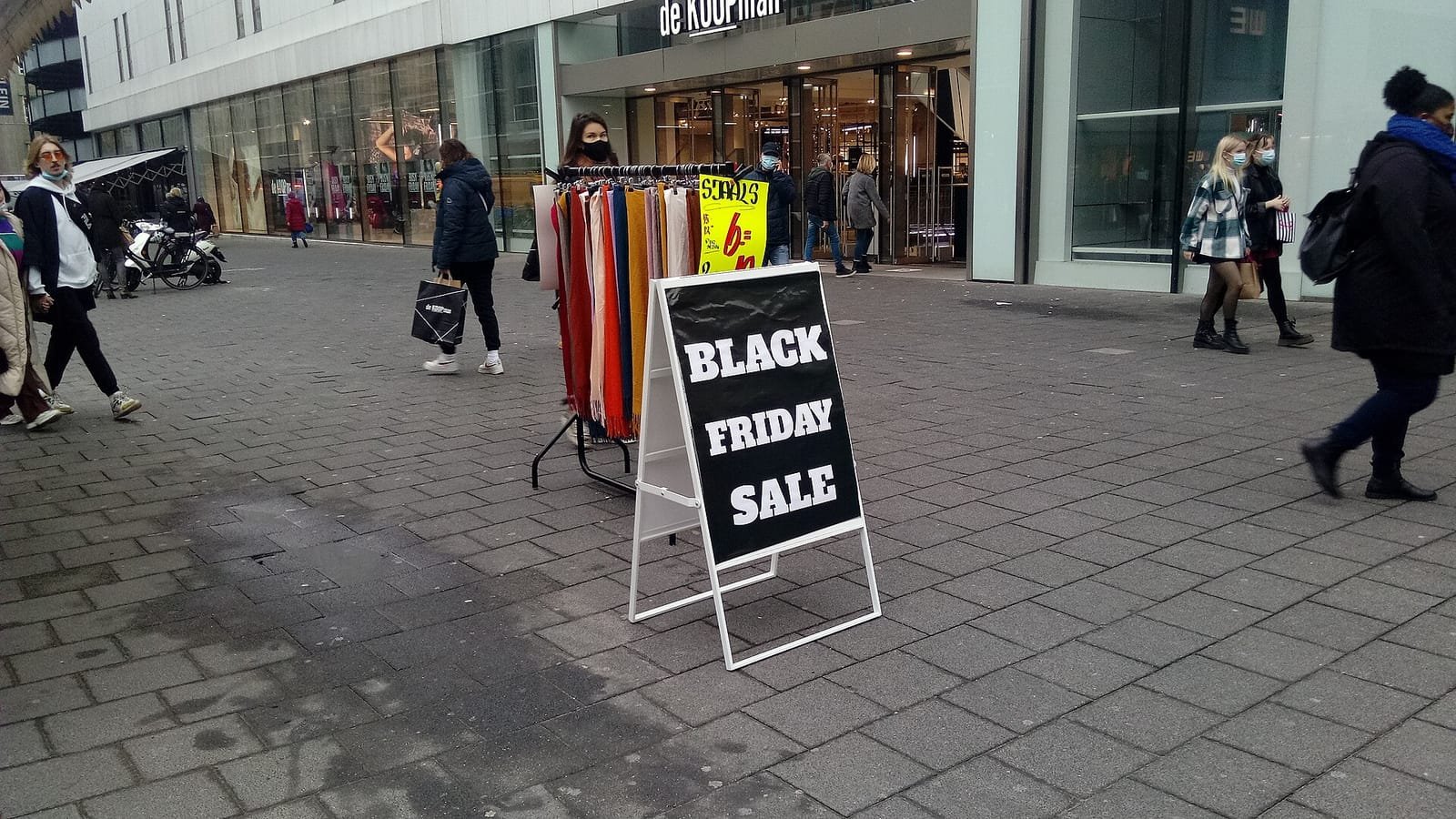 Cartel negro de «Black Friday Sale» en una calle peatonal de Róterdam, junto a un perchero con bufandas y varios peatones con mascarilla