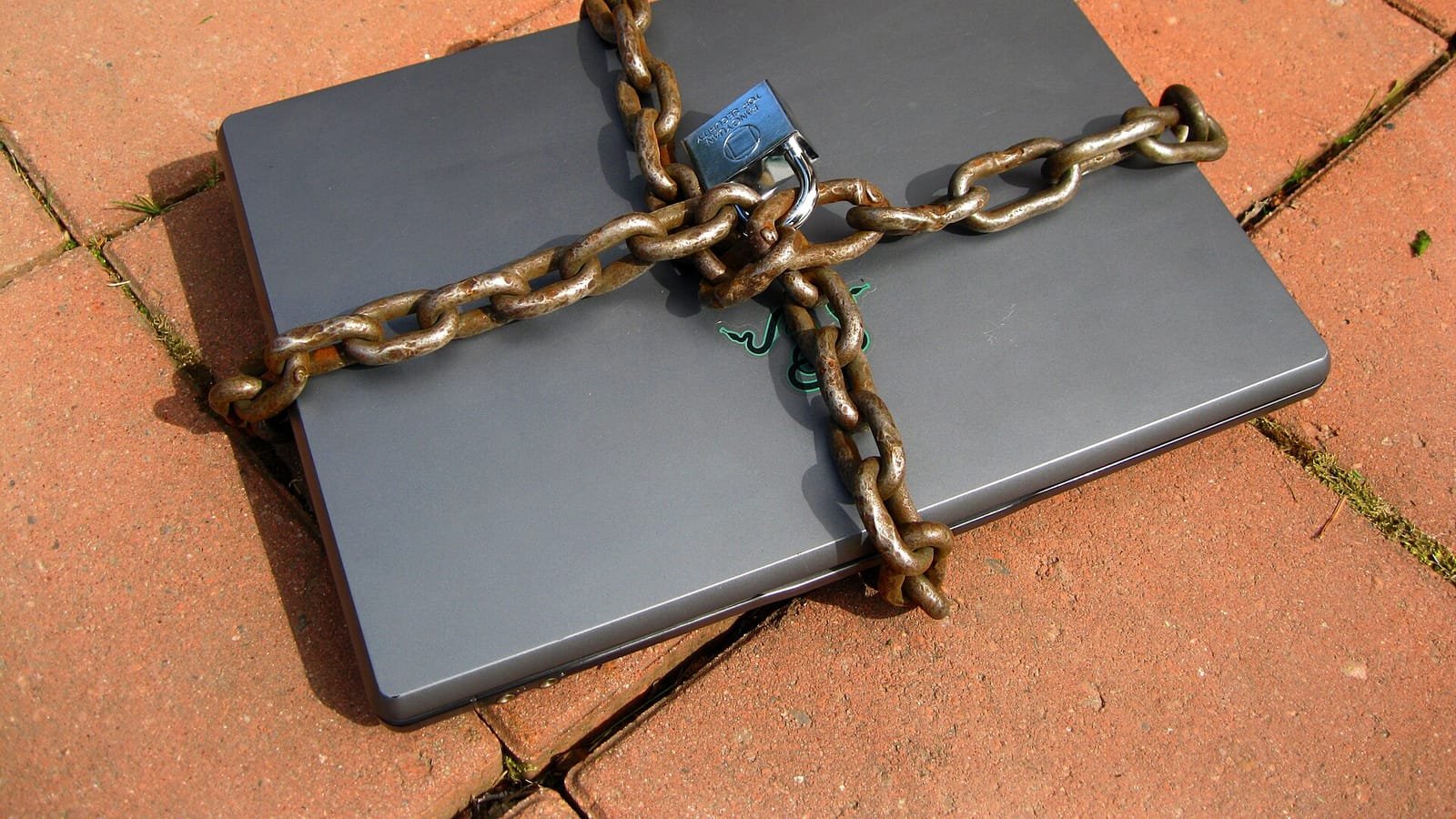 Un portátil gris cerrado y rodeado de cadenas oxidadas con un candado, sobre un suelo de baldosas de ladrillo